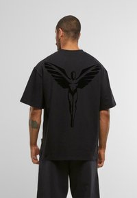 Mann iført svart oversized T-skjorte med stort svart vinget humanoid figurgrafikk på ryggen, som står mot en enkel lys grå bakgrunn.