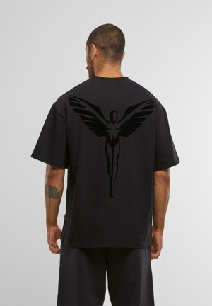 Vyras, vilkintis juodą per didelį T-shirt'ą su dideliu juodu sparnuotu humanoidu grafiku ant nugaros, stovi prieš pilką šviesią fono.