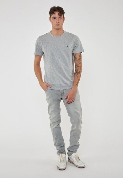 Maglietta in cotone grigia con maniche corte e un piccolo logo sul petto, abbinata a jeans blu chiaro e sneaker bianche.