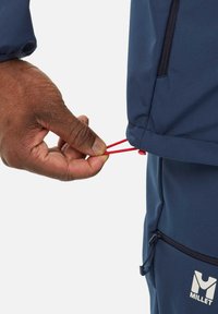 Veste extérieure marine avec cordon élastique ajustable. Main tenant un cordon rouge à l'ourlet, mettant en valeur un design slim et un tissu texturé.