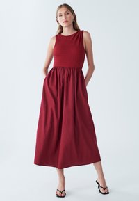 Rdeča maxi obleka brez rokavov z teksturiranim oprijetim zgornjim delom in nabranim krilom. Ima stranske žepe in gladko, tekočo silhueto.