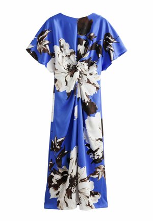 Frau trägt ein blaues, geblümtes Kleid mit kurzen Ärmeln und Schlitz vorne, hält eine schwarze Clutch und trägt schwarze Sandalen.