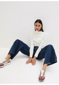 Haut blanc à manches longues, col montant ; jean en denim foncé à jambes larges ; baskets grises, bordeaux et blanches avec lacets et détails texturés.