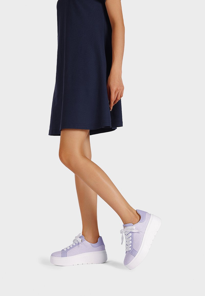 Sneakers viola e bianchi con tomaia texturizzata, suola bianca spessa e design con lacci. Indossati con un vestito blu navy, mostrano una vista laterale.