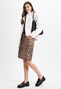 Weiße Jeansjacke über einem schwarzen Oberteil, kombiniert mit einem Bleistiftrock im Leopardenmuster. Schwarze Slipper vervollständigen das Outfit. Eine schlichte Handtasche in der Hand.