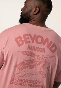 Růžové tričko s krátkým rukávem s nápisem "BEYOND MMXIII" a ilustrací okřídlené bytosti. Měkká, lehká látka.