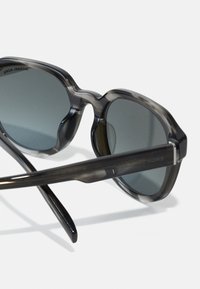 Ochelari de soare gri, semi-transparenți, cu lentile întunecate și un design unghiular. Prezintă un logo proeminent pe arma și o textură netedă pe întreaga suprafață.