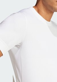 Camiseta deportiva blanca con una textura suave, cuello redondo y mangas cortas con paneles de malla en los laterales para mayor transpirabilidad.