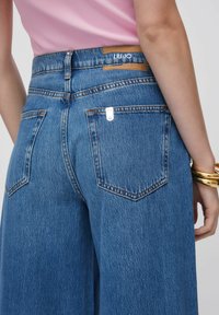 Jean en denim bleu à taille haute, doté de deux poches arrière et d'un patch en cuir avec le logo "LIU-JO". Texture lisse et coupe classique.
