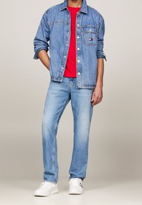 Man wearing light blue denim jacket and jeans, red shirt, and white sneakers, standing with one hand in pocket against plain background.