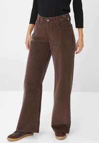Braune Cordhosen mit hoher Taille, weitem Bein, Fronttaschen und einem einzigen Metallknopfverschluss. Kombiniert mit dunklen Schuhen.
