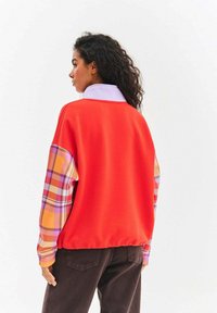 Czerwona bluza z tkaniną w kratę na rękawach w kolorach żółtym, purpurowym i pomarańczowym. Posiada biały kołnierz i luźny krój.