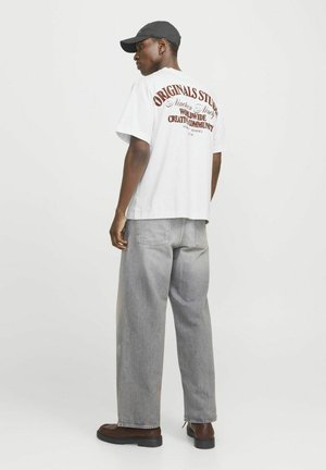 Witte t-shirt met korte mouwen en rode tekst op de achterkant, gecombineerd met lichtgrijze wijde jeans en bruine schoenen. Effen kleur pet gedragen.