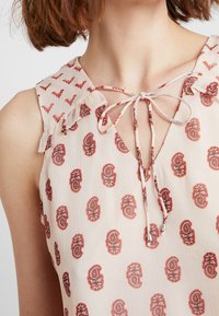 Blusa leggera di colore crema con stampa a paisley rossa. Presenta un colletto arricciato e un legaccio al collo con accenti di perline. Tessuto morbido e fluente.