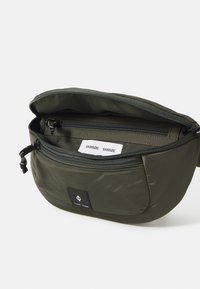 Borsa in nylon verde oliva con chiusura a zip, tasca interna e patch con logo nero. Presenta una texture liscia e una forma arrotondata.