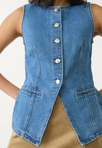 Die Jeansweste hat ein tailliertes Design, fünf silberne Knöpfe, zwei Seitentaschen und eine hellblaue Farbe mit sichtbaren Nähdetails.