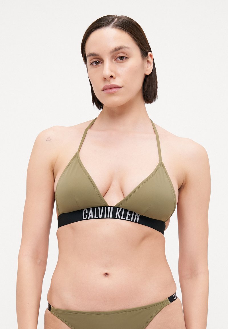Soutien-gorge triangle vert olive avec bande élastique noire portant le texte "CALVIN KLEIN". Les bretelles sont fines et le tissu semble lisse.