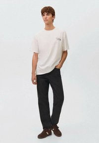 Camiseta blanca de algodón con un pequeño gráfico en el pecho, combinada con pantalones vaqueros negros y zapatillas marrones. Look simple y casual.