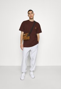 Bordo puuvillane T-särk, valged dressipüksid ja beežist crossbody-kott. Mudel valgete spordijalatsitega seisab neutraalses taustas.