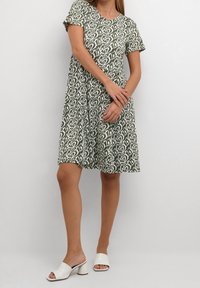 Zielona sukienka w wzory z krótkimi rękawami i okrągłym dekoltem, zdobiona białym kwiatowym wzorem. Połączona z białymi sandałami na słupku z otwartymi palcami.