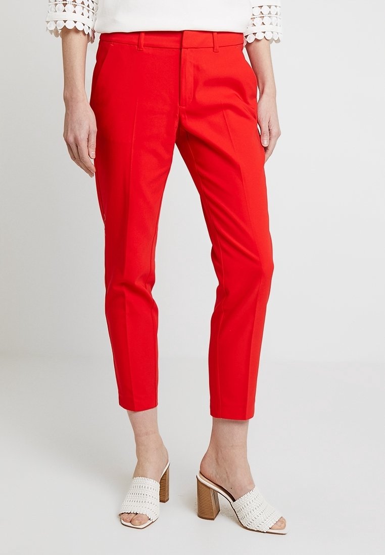 Pantalon rouge sur mesure à coupe ajustée, avec plis avant et texture lisse. Associé à des talons ouverts blancs tissés.