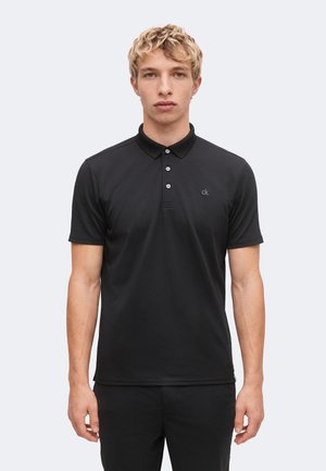 Jeune homme aux cheveux blonds courts et bouclés, portant un polo noir à manches courtes avec un petit logo "ck" sur la poitrine, debout devant un fond uni.