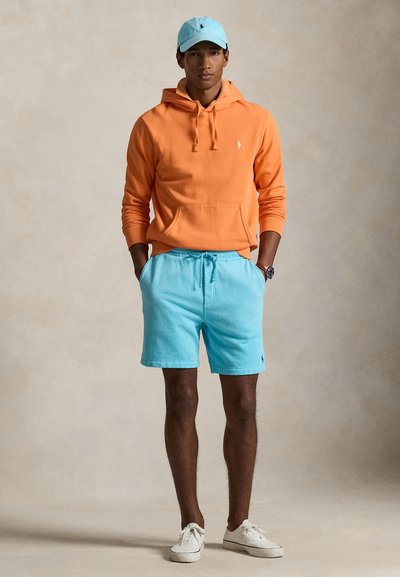 Homme portant un sweat à capuche orange, un short bleu clair, une casquette assortie, des baskets blanches et une montre au poignet, debout les mains dans les poches.