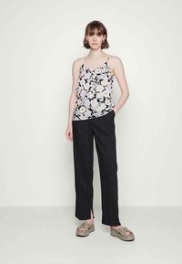 Jovem mulher de pé, vestindo uma blusa de alças finas em preto e branco com padrão floral, calças pretas com fendas frontais e sandálias de plataforma bege.