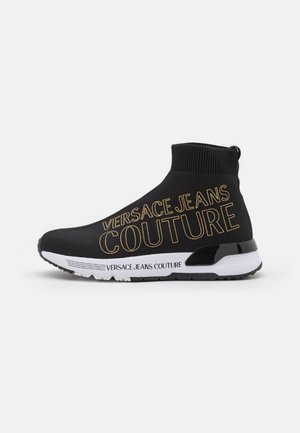 Czarny sneaker w stylu skarpetki z białą podeszwą i złotym napisem "Versace Jeans Couture" po boku i na podeszwie, z wysokim, dzianym kołnierzem nad kostką.