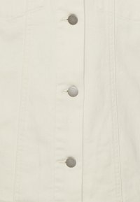 Chaqueta de mezclilla blanca con botones de metal plateado, dos bolsillos frontales y una tela suave y texturada con costuras visibles.