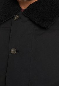 Detailní záběr na černý kabát s knoflíky a texturovaným fleece límcem nošený přes tmavou košili.