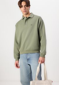 Polo de algodón verde con cuello y tapeta de botones, que presenta un logo bordado en el pecho. Llevado con jeans de color azul claro y una bolsa de mano beige.