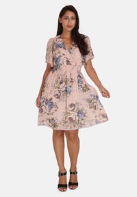 Blumenkleid in zartem Pink mit blauen und lila Rosenmustern. Kurze Ärmel, gesmokter Taillenbereich und knielanger Saum. Kombiniert mit schwarzen Schuhen.