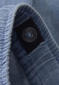Lichtblauwe denimstof met een zichtbare knoop en een zwart elastisch gedeelte. De stikseldetails benadrukken de textuur en vorm van het materiaal.