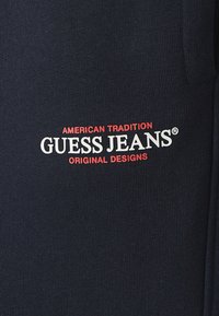 Tumši zilas kokvilnas audums ar rievotu tekstūru, centrā ir balta un sarkana "GUESS JEANS" logo un teksts "AMERICAN TRADITION".