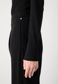 Pantaloni neri su misura con una texture liscia, caratterizzati da una piega laterale e un piccolo dettaglio metallico vicino alla vita. Camicia a maniche lunghe nera visibile.