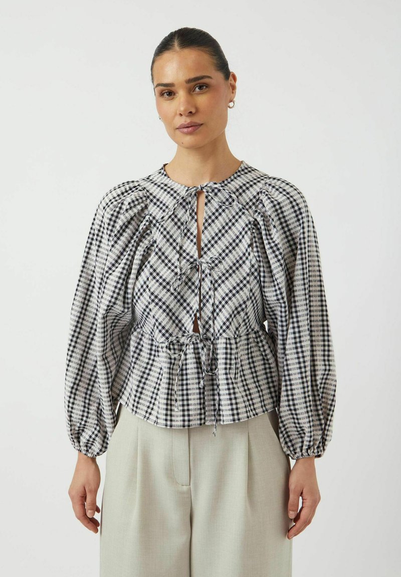 Blusa a scacchi in bianco e nero con maniche a sbuffo, dettagli arricciati, scollatura a chiave e vita regolabile con cordoncino. Abbinata a pantaloni chiari.
