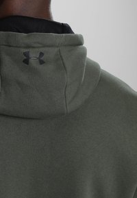 Sudadera con capucha verde que presenta un logo negro en la parte posterior del cuello. Textura suave con un ajuste relajado. Capa interior negra visible debajo de la sudadera.