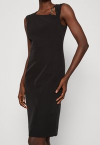 Schwarz ärmelloses Kleid mit quadratischem Ausschnitt, versehen mit einem goldfarbenen Akzent an der Schulter und einer taillierten Silhouette, gefertigt aus glattem Stoff.