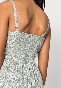 Vista trasera de una persona que lleva un vestido de lunares en verde y blanco, con tirantes finos ajustables y detalle de espaldas fruncidas.