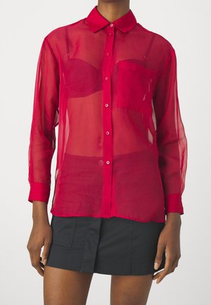 Osoba ubrana w przezroczystą czerwoną koszulę z guzikami na czerwonym biustonoszu oraz czarną mini spódniczkę, stojąca na jednolitym tle.