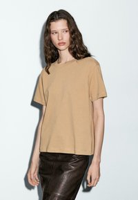 T-shirt in cotone color nocciola, maniche corte, colletto girocollo, vestibilità comoda, abbinato a una gonna in pelle marrone scuro, con texture opaca.