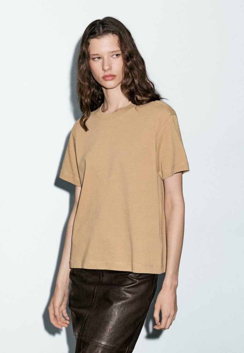 T-shirt in cotone color nocciola, maniche corte, colletto girocollo, vestibilità comoda, abbinato a una gonna in pelle marrone scuro, con texture opaca.