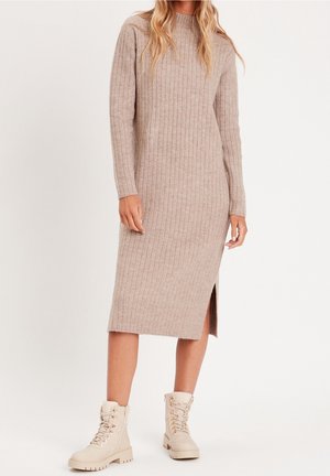 Femme portant une robe midi en maille côtelée beige avec manches longues et fente latérale, associée à des bottines beige clair à lacets.