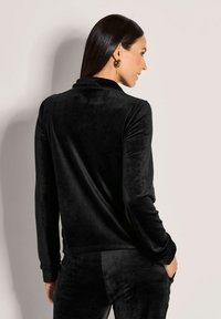 Vrouw met steil, donker haar, die gouden oorringen draagt en een zwarte, fluwelen top met lange mouwen en bijpassende broeken aanheeft, zijwaarts en achterwaarts kijkend.