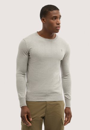 Uomo rivolto verso destra che indossa un maglione a maniche lunghe grigio chiaro e pantaloni cargo verde oliva su uno sfondo uniforme.