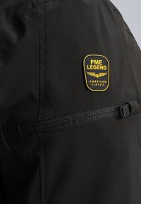 Schwarze Jacke mit einem PME Legend-Abzeichen in Gelb, mit einer Reißverschluss-Tasche und glatter Stofftextur. Sichtbare Nahtdetails.