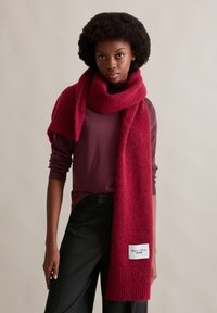 Dieprode wollen sjaal met een zachte, textuurrijke afwerking, losjes gedrapeerd. Heeft een label met "Marc O'Polo Denim" aan één uiteinde.