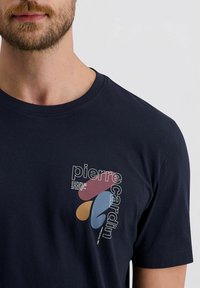 Man in een marineblauwe t-shirt met abstracte vormen en de tekst "pierre cardin" gedrukt op de linkerkant van de borst.