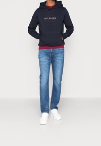 Felpa blu con logo, accenti rossi, tasca frontale; abbinata a jeans azzurri chiari e sneakers bianche. Outfit semplice e casual.